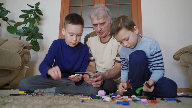 Two little brothers and grandfathers are sitting on the floor and assembling parts of the constructor for the construction of various buildings in living room. blocks.