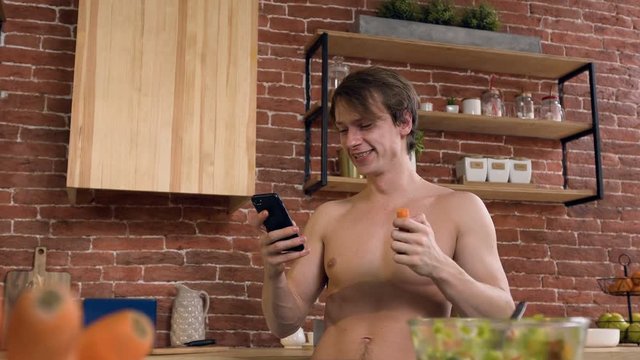 Attractive sport man eating carrot while surfing in internet on smart phone at home.