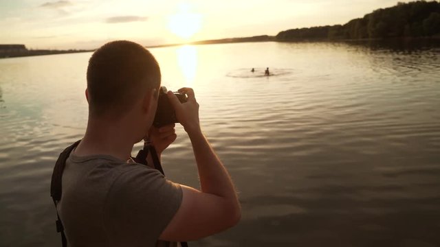 A man is standing with his back and photographing the sunset on the lake surrounded by trees on the background with bathing people.