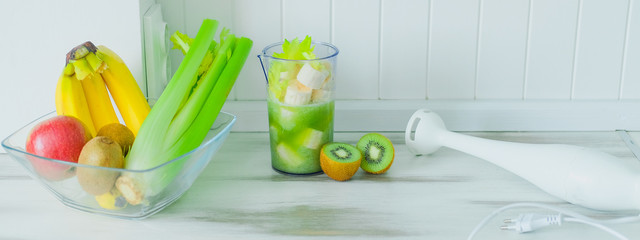 Green smoothie of celery and banana. Cut ingredients and white immersion blender on table. Healthy...