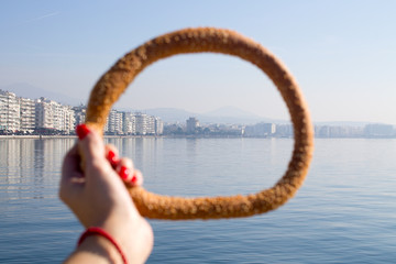 womans hand with red maniqure holding the greek bagel koulouri with sea and city on the background