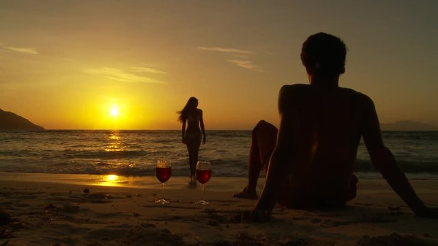 WS Woman walking towards man sitting with glasses of wine on beach at sunset / Seychelles
