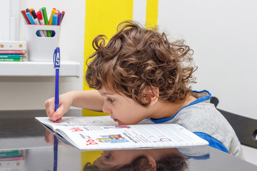 Tired little boy sitting at the table, writing in her notebook while doing homework. Concept of a large number of assignments at school