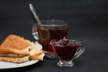 Tea with raspberry jam and pancakes. Cup of transparent glass, sauce-boat and white plate. Dark food photo style.