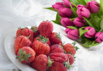 Beautiful and delicious strawberry on a white plate, near a bouquet of pink tulips