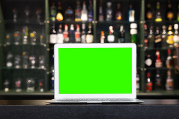 Blank screen of Laptop on blur alcohol drink bottle on bar counter in the dark night background.