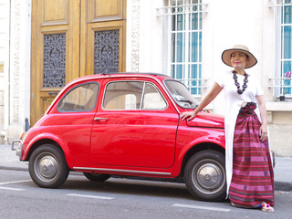Femme avec jupe longue appuyée sur une ancienne fiat 500 rouge.