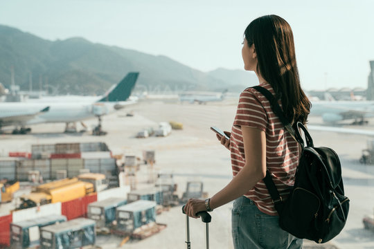 young girl seeing view of busy airplanes apron standing near window holding cellphone backpack and luggage in hall walking to lounge area. travel woman search online map in hong kong airport to gate