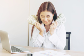 Successful beautiful Asian business young woman holding money US dollar bills in hand on office desk, business concept