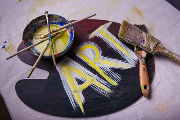 Word ART painted in white and yellow acrylic paint on paint mixing pallet paintbruses scattered and stacked  