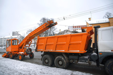 Orange snow removal machine loads snow into a dump truck for removal from city. Emergency cleans road after heavy snowfall  by municipal workers. Road cleaning with special equipment vehicle