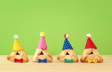 Purim celebration concept (jewish carnival holiday). Traditional hamantaschen cookies with cute clown hats over wooden table and green background.