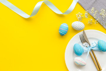 Easter table decoration. Plate, cutlery, painted eggs and dry white flowers, tablecloth on yellow background top view copy space