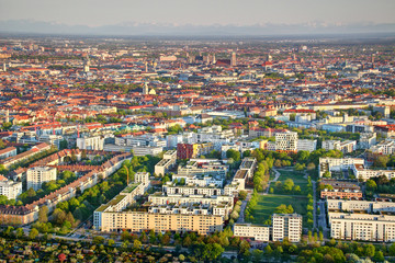 Aerial view of city outskirts and historic center with sunlit apartment buildings, housing estates, green parks, church towers and snowy Bavarian Alps in background Munchen Bayern Germany Europe
