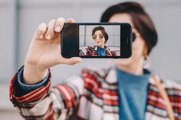 Close-up portrait of pretty and modern young fashion guru taking a selfie with her mobile phone,...