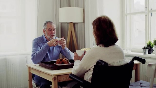 Senior couple with wheelchair sitting at the table at home, having breakfast and talking.