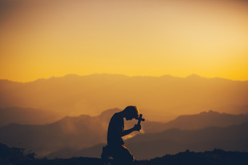 Young man kneeling down praying and holding christian cross for worshipping God at sunset...