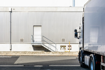 A white semitrailer truck parked opposite and perpendicularly to an industrial building with a pedestrian doorway at the top of a small staircase and covered with a grey corrugated iron siding. - obrazy, fototapety, plakaty