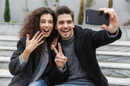 Image of european couple man and woman 20s in warm clothes, taking selfie photo on cell phone while sitting on stairs outdoor