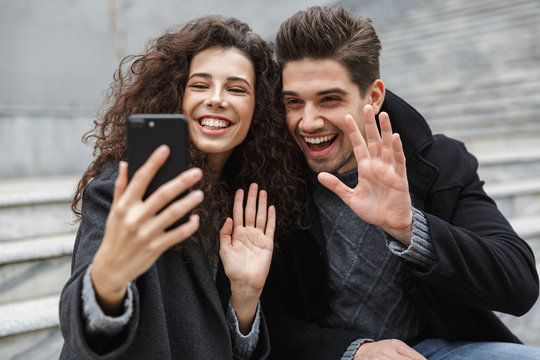 Image of happy couple man and woman 20s in warm clothes, taking selfie photo on cell phone while sitting on stairs outdoor - Powered by Adobe
