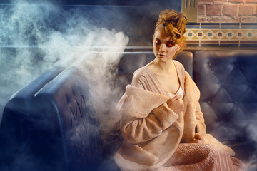 Glamor girl in stylish clothes and with a make-up sits on a leather sofa in the interior of the bar against the background of smoke.