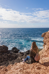 Fototapeta na wymiar Happy female tourist in coat sits on cliff and enjoys sea view in Malta