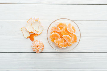 top view of peeled tangerine slices in glass bowl and peel on wooden white background