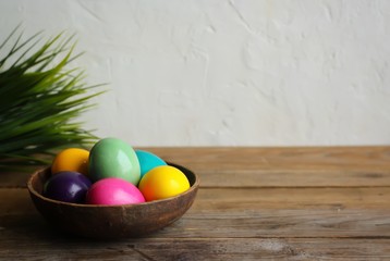 Colorful easter eggs in a brown bowl and grass. Wooden table, light background. Copy space, horizontal orientation