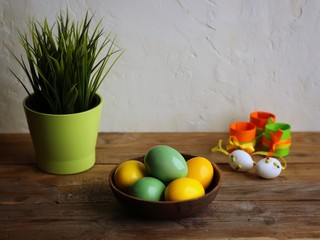 Colorful easter eggs in a brown bowl, grass in a green flowerpot. Wooden table, light background. Copy space, horizontal orientation