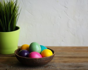 Colorful easter eggs in a brown bowl, grass in a green flowerpot. Wooden table, light background. Copy space, horizontal orientation