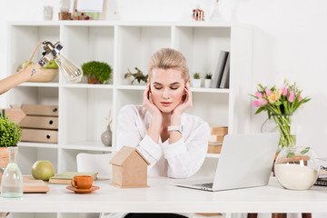 attractive woman looking at small cardboard house while sitting near laptop in office, environmental saving concept