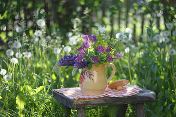 Still life in rustic style on a sunlit lawn with a bunch of summer flowers in a jug on a small bench