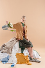 blonde woman sitting on stack of clothing and holding watering can with flowers on beige background, environmental saving concept