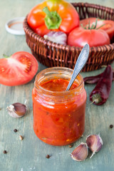 A glass jar with freshly prepared Georgian adjika sauce, a red ripe tomatoes, pepper and garlic on an old wooden table. Selective focus.
