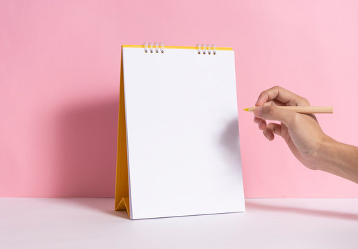 Hand holding pencil for writing marking on mockup paper spiral calendar on pink background.