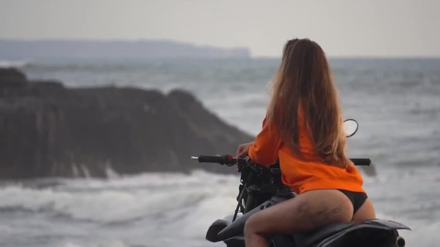 wonderful back shot of a young model in orange hoodie posing on a motorbike, looks at the ocean with big waves splashing against the rocks,