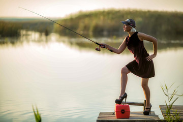 Pretty woman in a burgundy dress and black shoes and a baseball cap catches fish on a wooden pier...
