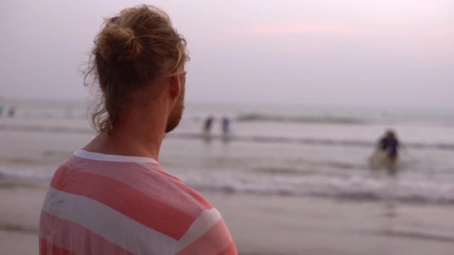 Slowmo back side view handsome relaxed dreamy blond bearded man hair combed bun, contemplating landscape during vacation, tourist watching ocean, sufer conquer waves, enjoying travel warm weather