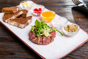 Beef tartare with arugula salad, crisp bread chips, sauces and snacks on white plate on the served restaurant table.
