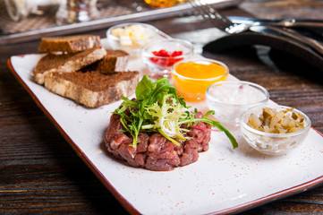 Beef tartare with arugula salad, crisp bread chips, sauces and snacks on white plate on the served restaurant table.