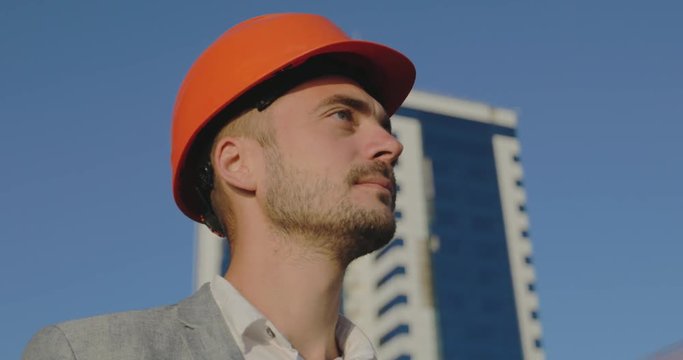 close view engineer with designer stubble in orange helmet looks into distance against high building under blue sky