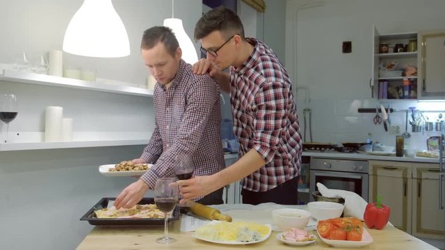 Couple of men gay flirts each other cooking a pizza together and drinking a wine.