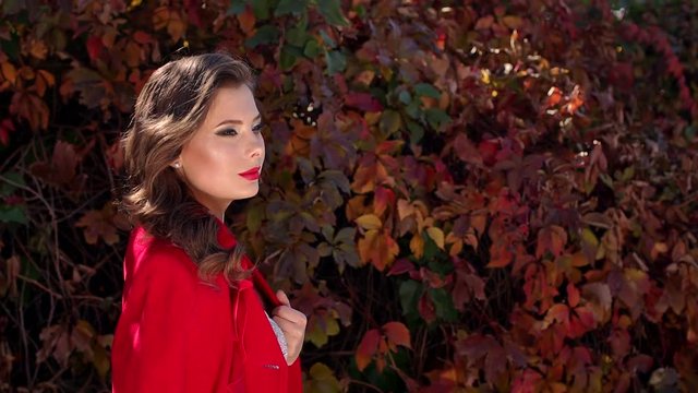Close-up of a beautiful girl with bright make-up, hairstyle and a red coat walking in autumn park against the background of bright colored autumn leaves.