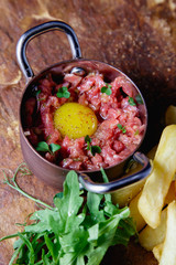 Carpaccio made of chopped tender veal with raw chicken eggs, served in a small metal pot on a stone table and a small amount of fries and fresh herbs.