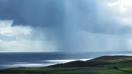 Heavy rain over calm ocean in the background. Green cliff in foreground.