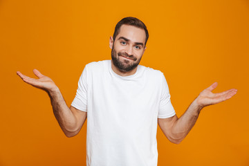 Image of puzzled man 30s in t-shirt throwing up hands, isolated over yellow background