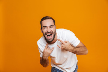 Image of happy guy 30s in t-shirt rejoicing and showing rock sign while standing, isolated over yellow background