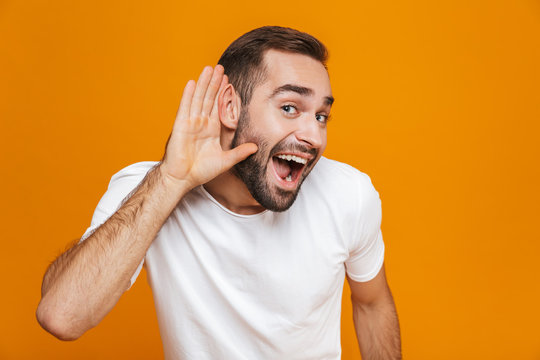 Image of joyful man 30s trying to hear something while keeping hand at his ear, isolated over yellow background