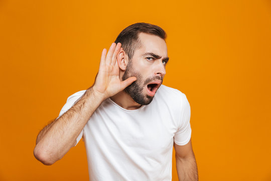 Image of european man 30s trying to hear something while keeping hand at his ear, isolated over yellow background