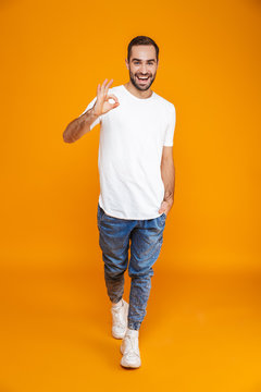 Full length image of handsome guy 30s in t-shirt and jeans showing ok sign while standing, isolated over yellow background